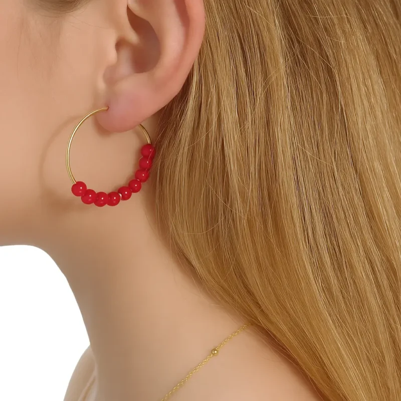 pendientes tipo criolla con un fino aro de metal dorado. La mitad inferior de cada pendiente está decorada con una secuencia de cuentas esféricas de color rojo brillante, interrumpida por dos pequeñas cuentas doradas.