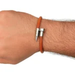 Una pulsera de cuero liso de color marrón anaranjado con un cierre y detalles de metal plateado en forma de clavo, sobre un fondo blanco.